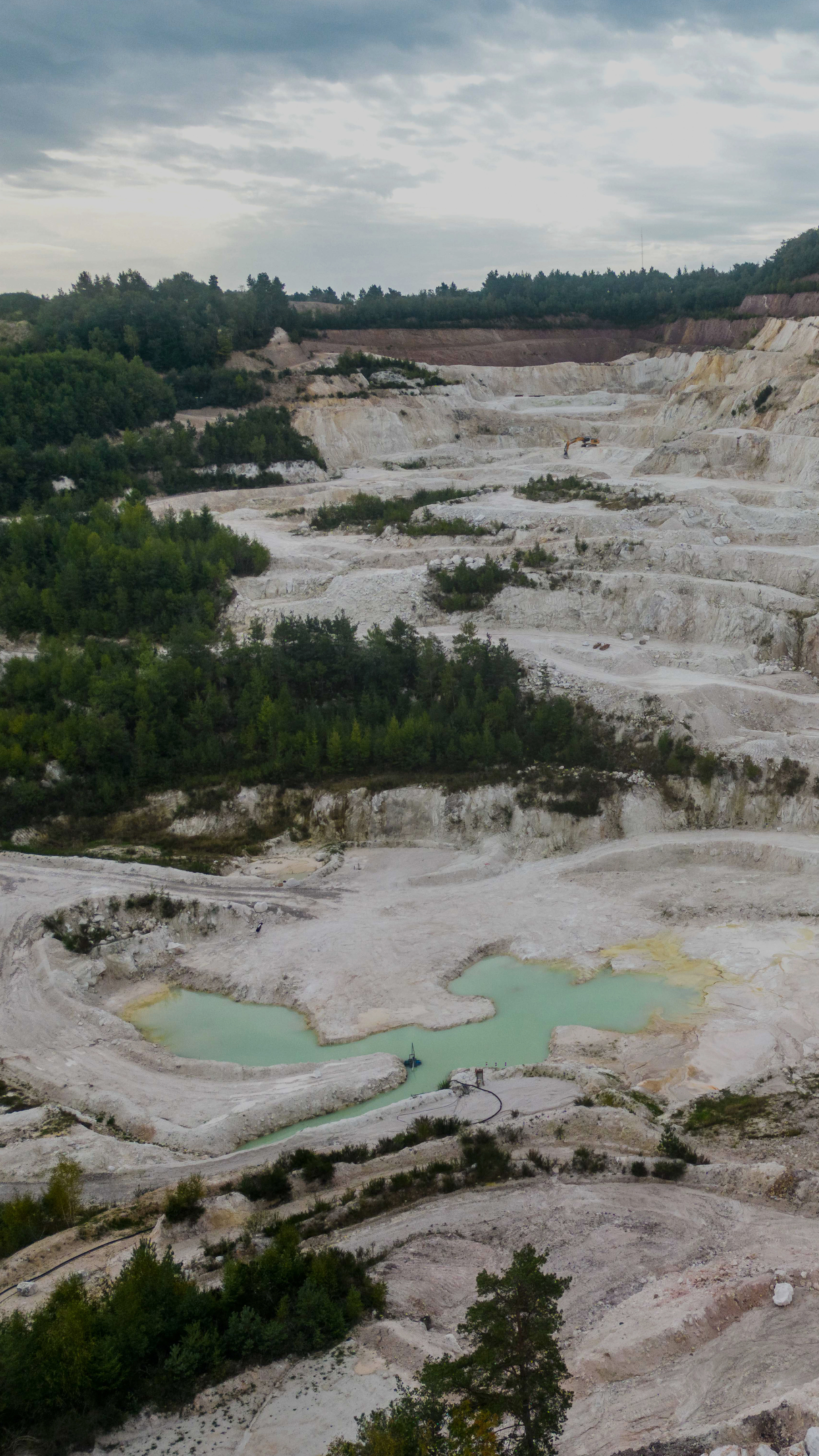 Carrière à ciel ouvert avec des bassins d’eau turquoise, des strates rocheuses exposées, entourée de végétation et de collines boisées.