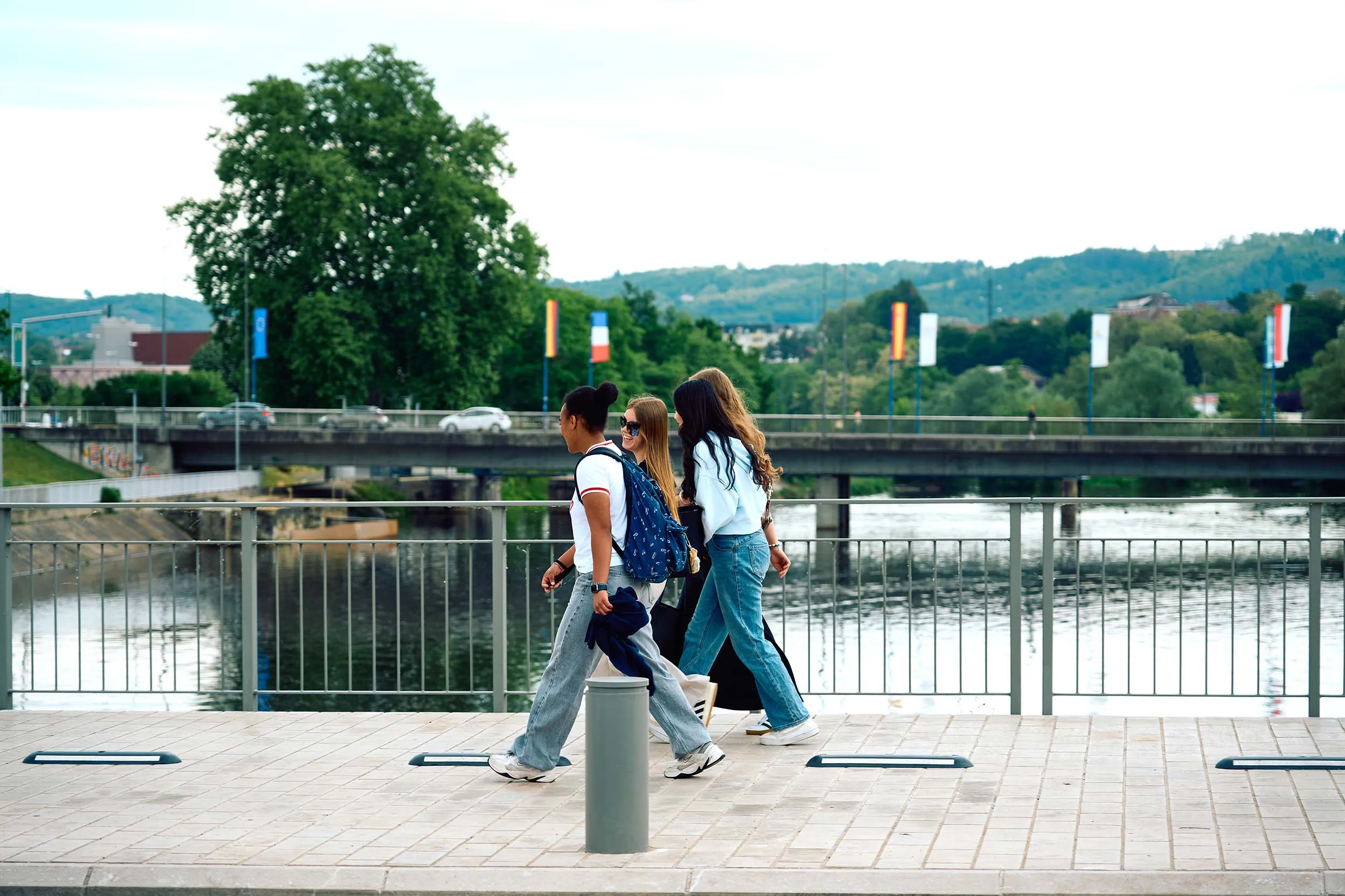 Trois jeunes marchent sur un trottoir en bord de rivière, un pont et des collines verdoyantes en arrière-plan.