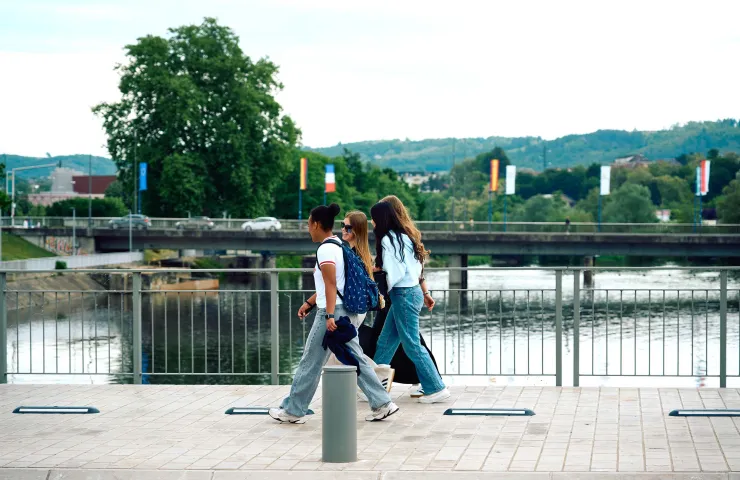 Trois jeunes marchent sur un trottoir en bord de rivière, un pont et des collines verdoyantes en arrière-plan.