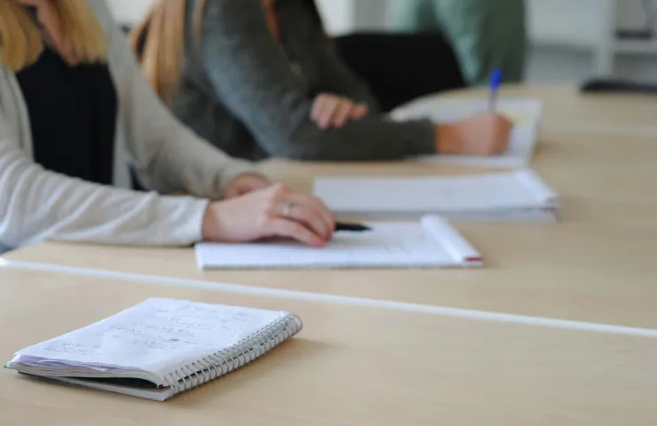 Personnes en réunion prenant des notes manuscrites sur cahiers, assises à une table en bois dans un environnement professionnel